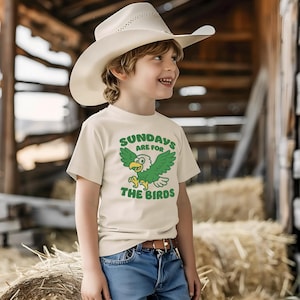 Op de afbeelding: Een cr&egrave;mekleurig T-shirt met de tekst "SUNDAYS ARE FOR THE BIRDS" en een groene cartoon adelaar. Het kind draagt een witte cowboyhoed en een blauwe spijkerbroek. Het shirt heeft een bruine riem.