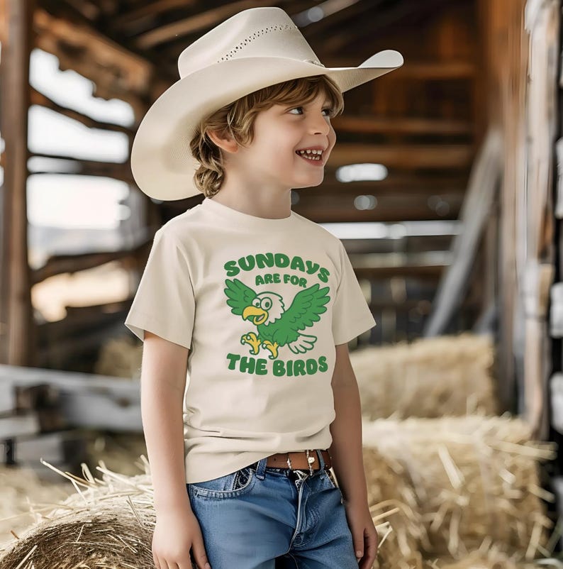 Op de afbeelding: Een cr&egrave;mekleurig T-shirt met de tekst "SUNDAYS ARE FOR THE BIRDS" en een groene cartoon adelaar. Het kind draagt een witte cowboyhoed en een blauwe spijkerbroek. Het shirt heeft een bruine riem.
