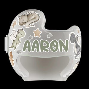 Puede incluir: Casco de beb&eacute; transparente y curvado, decorado con ilustraciones de dragones, estrellas y el nombre "AARON" en verde. El dise&ntilde;o del casco es principalmente gris y blanco, creando una est&eacute;tica suave y juguetona.