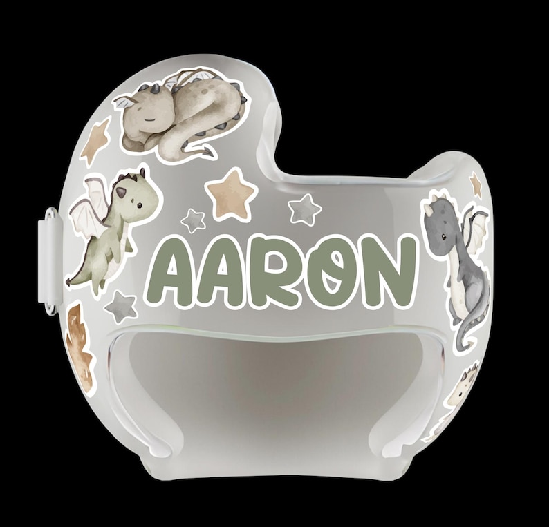Puede incluir: Casco de beb&eacute; transparente y curvado, decorado con ilustraciones de dragones, estrellas y el nombre "AARON" en verde. El dise&ntilde;o del casco es principalmente gris y blanco, creando una est&eacute;tica suave y juguetona.