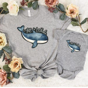 Op de afbeelding: Een grijs T-shirt en een grijs rompertje met een blauwe walvisafbeelding en de tekst "All is Whale".