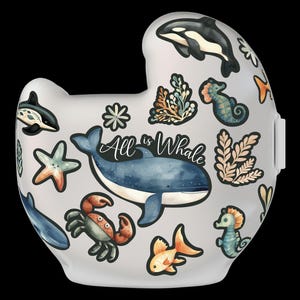 Op de afbeelding: Een witte keramische mok met een fantasierijk ontwerp met een oceaan thema. De mok heeft een blauwe walvis, een zeester, een krab, zeepaardjes en zeewier. De tekst "All is Whale" is geschreven in zwarte cursieve letters.