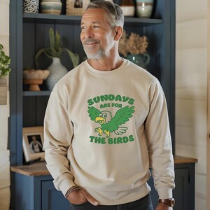 Op de afbeelding: Beige sweatshirt met de tekst "SUNDAYS ARE FOR THE BIRDS" en een groene cartoon adelaar. Het sweatshirt met lange mouwen wordt gedragen door een persoon voor een blauwe kast. De tekst en afbeelding zijn groen.