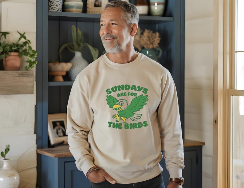 Op de afbeelding: Beige sweatshirt met de tekst "SUNDAYS ARE FOR THE BIRDS" en een groene cartoon adelaar. Het sweatshirt met lange mouwen wordt gedragen door een persoon voor een blauwe kast. De tekst en afbeelding zijn groen.