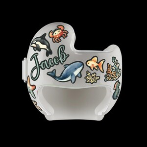 Op de afbeelding: Een witte helm met stickers van een blauwe walvis, een oranje vis, een krab en zeewier. De helm heeft de naam "Jacob" erop.