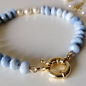 May include: A beaded bracelet featuring light blue and white faceted beads, transitioning to white pearls. The bracelet is secured with a gold-colored clasp. Additional gold-colored jewelry is visible in the background.