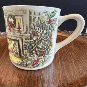Puede incluir: Taza de cerámica color crema con un diseño navideño. La ilustración presenta un árbol de Navidad decorado, una chimenea y una corona. La taza tiene un asa curva. El fondo es marrón.