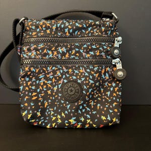 May include: Black crossbody bag with a colorful bird pattern. The bag has three zippered compartments, a black adjustable strap, and a round logo patch. The bag's pattern includes blue, orange, yellow, and purple birds.