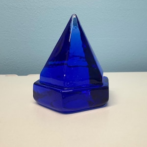 May include: A cobalt blue glass paperweight shaped like a pyramid with a hexagonal base. The object is made of solid, translucent blue glass with smooth, angled surfaces that meet at a sharp point at the top, reflecting light across its facets.