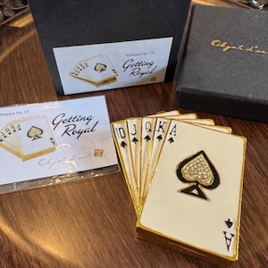 May include: A decorative box shaped like a fan of five playing cards, featuring an ace of spades with a crystal-encrusted spade symbol. The set includes a black storage box and a card labeled Release No. 13 Getting Royal with a matching illustration.