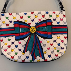 May include: White canvas shoulder bag with a colorful heart pattern and a large bow design. The bow features red, blue, and green stripes, with the words "Joy Love" in the center. The bag has a black strap.
