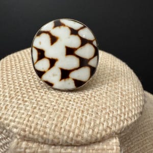 May include: A circular ring featuring a white and brown pattern. The ring has a silver-coloured band and a large, round face. The brown pattern consists of irregular shapes set against a white backdrop.