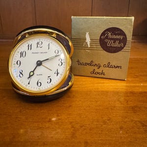 May include: A vintage Phinney-Walker traveling alarm clock with a gold-tone bezel and a white face. The clock has black hands and numerals. The original box is included, with the words "Phinney-Walker" and "traveling alarm clock" printed on it.
