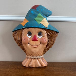 May include: A molded scarecrow head wall decoration featuring a blue hat with orange, green, and yellow diamond patches. The face has a red triangular nose, brown hair, and a rope-like collar detail. The surface shows signs of paint wear.