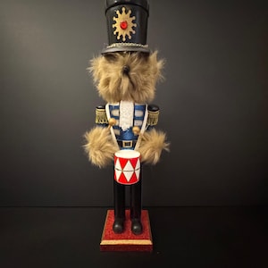 May include: A decorative nutcracker in the shape of a teddy bear soldier. The bear wears a blue and white uniform with gold accents, a black hat with a gold sunburst design, and holds a red and white drum. The base is red.