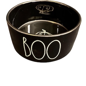 Puede incluir: Cuenco de cerámica negro con borde plateado y la palabra "BOO" en letras blancas. El cuenco tiene un diseño de calabaza en el interior, ideal para decoraciones de Halloween.