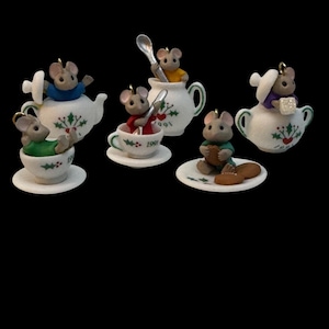 May include: A collection of five miniature mouse figurines, each interacting with white ceramic teaware decorated with holly leaves and berries. The mice wear colorful shirts and are positioned in or near teapots, cups, and saucers, with some holding spoons.