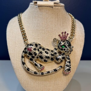 May include: Gold-tone necklace featuring a bejeweled leopard pendant with black spots, green eyes, and a pink crown. The leopard has pink accents on its paws and mouth. The necklace is displayed on a beige bust.