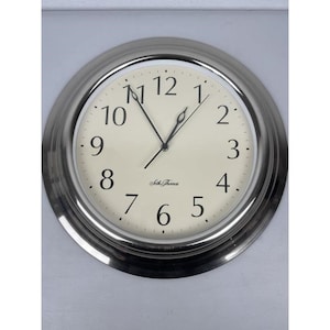 Peut inclure: Une horloge murale ronde, argentée, avec un cadran crème. Des chiffres noirs indiquent les heures, et des aiguilles noires indiquent l'heure. Le cadran porte le nom de la marque "Seth Thomas" en noir.