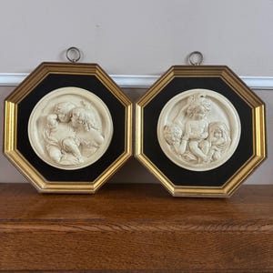 May include: Two framed relief sculptures. Each features a circular, off-white bas-relief scene of cherubic figures. The frames are octagonal with a black inner border and gold outer frame. Each has a small metal ring for hanging.