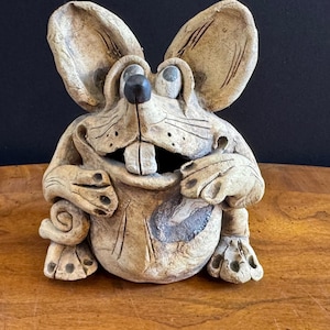 May include: A whimsical ceramic mouse sculpture with large ears, a black nose, and prominent teeth. The sculpture has a textured, beige finish with dark accents and is posed in a seated position. The mouse's expression is playful, making it a unique decorative item.