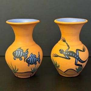 May include: Two terracotta vases with a light blue interior. One vase features a black and blue fish design, and the other has a black and blue lizard design. The vases are painted with orange and blue accents.