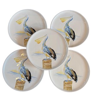 May include: A set of five white ceramic plates, each with a rope-style edge. The plates display a watercolour painting of a pelican on a wooden post, with a yellow beak and blue and brown plumage. The background shows a sunset over the sea.