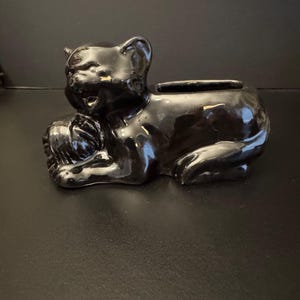Puede incluir: Figura de gato de cerámica negra brillante, tumbada con las patas cruzadas. El gato tiene una pequeña abertura en la espalda, posiblemente para una pequeña planta o un objeto decorativo. La figura es de color oscuro sólido.