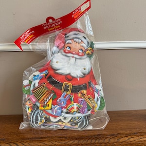 Könnte beinhalten: Ein Vintage-Adventskalender vom Weihnachtsmann in einer durchsichtigen Plastiktüte. Der Kalender zeigt einen großen, lächelnden Weihnachtsmann mit rotem Anzug, weißem Bart und schwarzem Gürtel. Der Kalender hat ein rotes Banner mit der Aufschrift "Adventskalender Santa Claus".