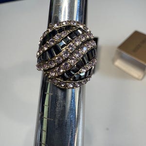 Könnte beinhalten: Ein Statement-Ring mit einem großen, abgerundeten Design. Der Ring hat abwechselnde Reihen aus schwarzen rechteckigen Steinen und kleinen, runden, hellvioletten Steinen. Der Ring ist auf einem silbernen Ringmaß angebracht.
