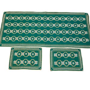 May include: A green embroidered table runner and two matching placemats. The set features a repeating pattern of white and gold floral designs. The edges are trimmed with a gold border. The table runner measures approximately 152 cm long.