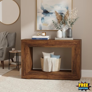Op de afbeelding: Een donkerbruine houten console met een rechthoekig frame. De tafel is versierd met boeken, kaarsen en vazen. Een gevlochten mand met kussens en een deken staat op de onderste plank. Een schilderij en een spiegel staan op de achtergrond.