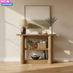 May include: A light wood console table with two shelves. The top shelf holds a lamp, books, and candles. The lower shelves display baskets, a vase, and a decorative bowl. A framed artwork hangs above the table.