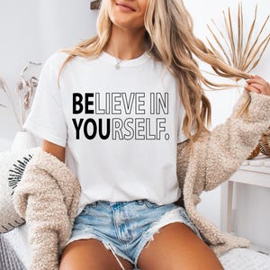 Pu&ograve; includere: Una maglietta bianca con la scritta "BELIEVE IN YOURSELF." stampata in nero.