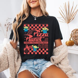 May include: Black t-shirt with a graphic of the Pizza Planet restaurant from the Toy Story movie. The graphic features the restaurant's logo, a checkered pattern, and the text "Pizza Planet", "Friday Night Kids Night", and "Get 1 Free Pizza when you order 2 Large Pizzas".