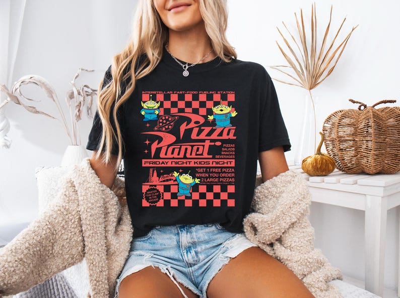 May include: Black t-shirt with a graphic of the Pizza Planet restaurant from the Toy Story movie. The graphic features the restaurant's logo, a checkered pattern, and the text "Pizza Planet", "Friday Night Kids Night", and "Get 1 Free Pizza when you order 2 Large Pizzas".