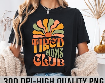 May include: A black t-shirt with a retro-style graphic design featuring a sun with a heart in the center and the text "Tired Moms Club".