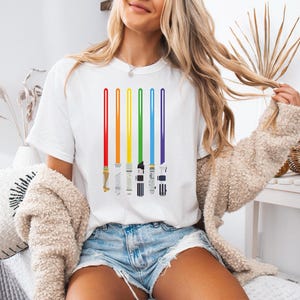 Peut inclure: T-shirt blanc avec un motif de sabres laser arc-en-ciel. Les sabres laser sont repr&eacute;sent&eacute;s en rouge, orange, jaune, vert, bleu et violet. Le t-shirt a un col rond et est fait d'une mati&egrave;re douce.
