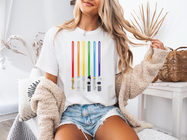 Peut inclure: T-shirt blanc avec un motif de sabres laser arc-en-ciel. Les sabres laser sont repr&eacute;sent&eacute;s en rouge, orange, jaune, vert, bleu et violet. Le t-shirt a un col rond et est fait d'une mati&egrave;re douce.