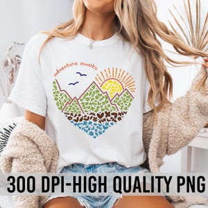 May include: White t-shirt featuring a colourful graphic design of mountains, a sun, and birds. The text "adventure awaits" is written above the graphic.
