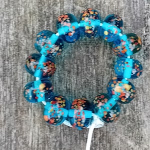 Puede incluir: Un brazalete circular de cuentas con grandes cuentas azules translúcidas. Las cuentas tienen patrones moteados en naranja, amarillo y negro. Las cuentas están ensartadas con un cordón azul claro, creando un accesorio vibrante.
