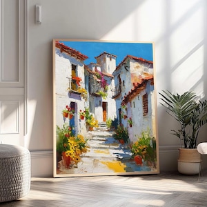 Op de afbeelding: Een ingelijst olieverfschilderij toont een zonnige Europese straat. Witte gebouwen met rode pannendaken flankeren een geplaveide weg, versierd met bloempotten. Het kunstwerk heeft een helderblauwe lucht en warme kleuren.