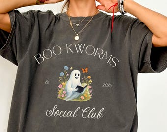May include: Dark gray t-shirt with the words "BOO-KWORMS Social Club" and a graphic of a friendly ghost reading a book, surrounded by flowers and butterflies. The shirt also has the text "Est 2025".