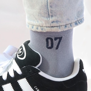 Op de afbeelding: Zwarte suède Adidas Campus sneakers met witte strepen en een witte zool. De schoenen worden gedragen met lichtblauwe jeans en grijze sokken met daarop "07".