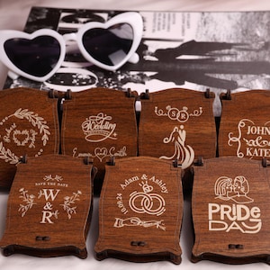 Puede incluir: Una colección de cajas de madera con varios diseños y textos grabados, incluyendo temas de boda y Orgullo. Gafas de sol blancas en forma de corazón descansan encima. Las cajas son marrones con grabados claros.
