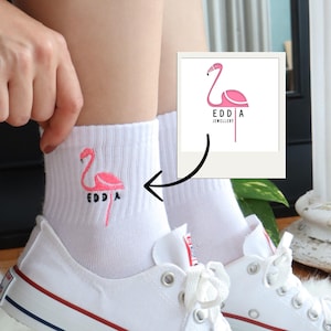 Könnte beinhalten: Weiße Knöchelsocken mit einem gestickten Design eines rosa Flamingos und dem Text "EDDA". Die Socken werden mit weißen Leinenschuhen mit roten und blauen Akzenten getragen.