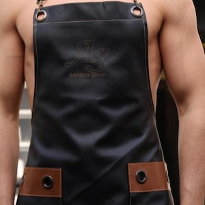 May include: A black leather apron with brown leather accents and the text "AS BARBER SHOP" embroidered on the front.