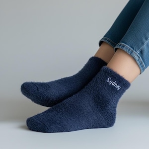 Peut inclure: Une paire de chaussettes duveteuses bleu marine avec le nom "Sydney" brodé en blanc sur le bord. Les chaussettes sont portées avec un jean bleu. Les chaussettes semblent douces et confortables.