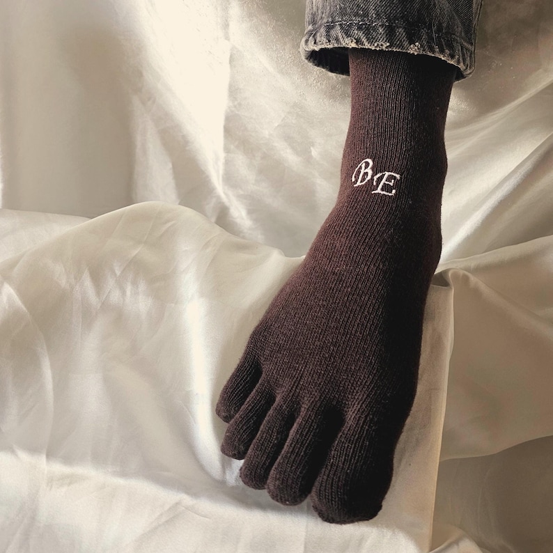 May include: A dark brown toe sock with the embroidered letters "BE" in white. The sock is made of a ribbed knit material and covers the foot and toes separately. The sock is worn with denim jeans.