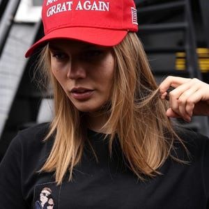 Puede incluir: Gorra de b&eacute;isbol roja con el texto blanco "MAKE AMERICA GRRAT AGAIN". La gorra tiene una visera curva y un peque&ntilde;o dise&ntilde;o de la bandera estadounidense en el lateral. La persona lleva una camiseta negra con un peque&ntilde;o parche bordado.